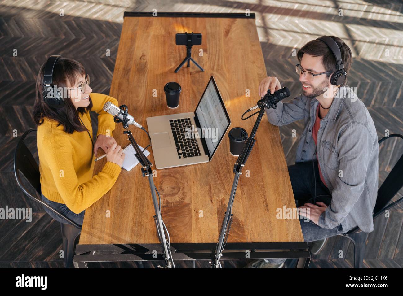 Two colleagues podcasters in headphones and glasses shooting video podcast using smartphone on tripod. Woman host blogger recording content with her guest in studio, interviewing, making notes. Stock Photo