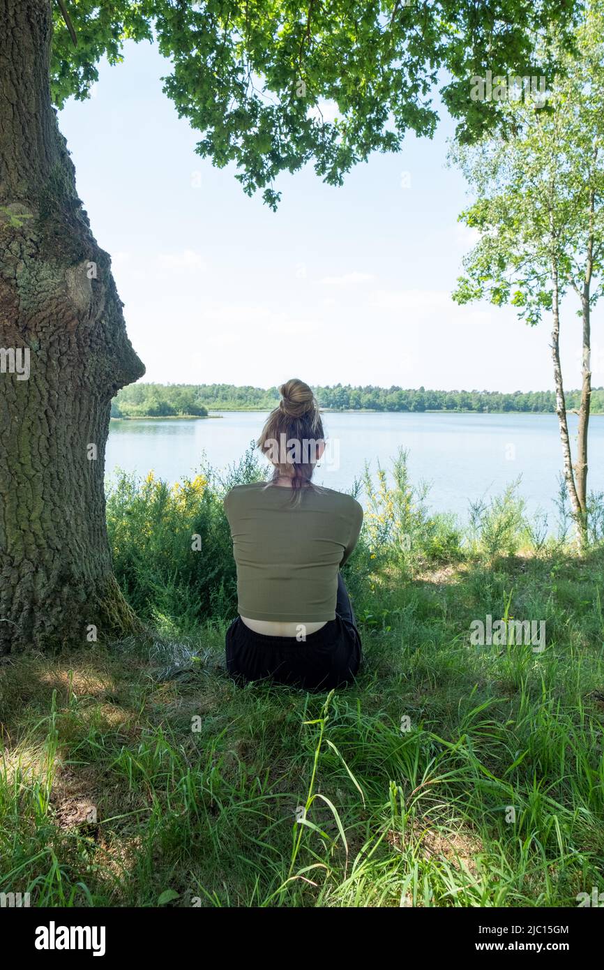 Young brunette woman sitting in green forest enjoys the silence and beauty of nature watching over a blue forest lake on a summer day. High quality photo Stock Photo