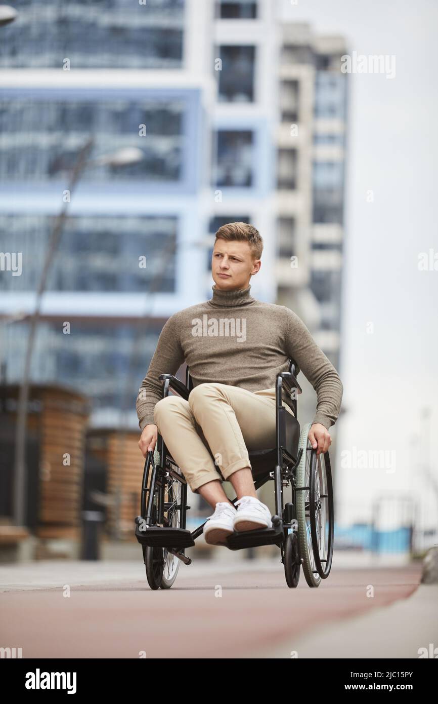 Serious pensive young man in casual sweater turning handrails while using wheelchair outside Stock Photo