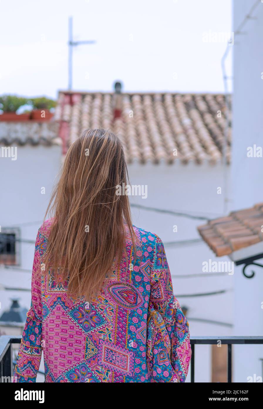 blonde woman seen from behind on balcony overlooking rural village street Stock Photo