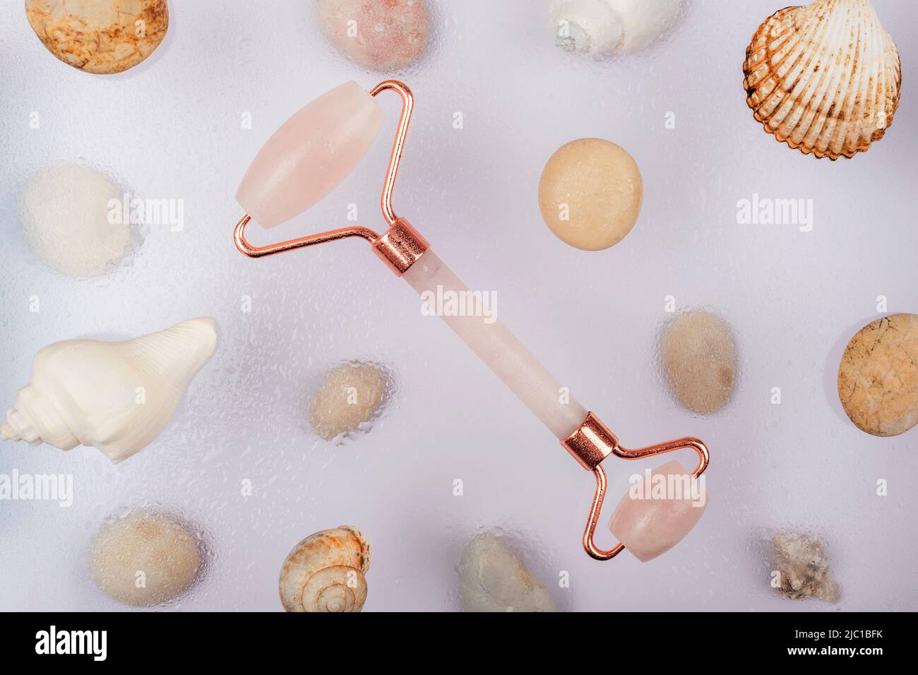 Rose quartz face roller and sea shells with water drops on white background. Selfcare concept. Top view, flat lay. Stock Photo