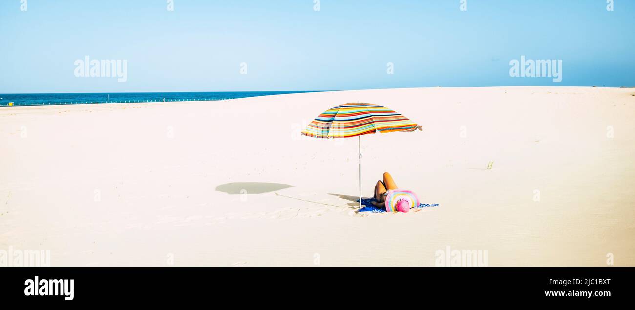 Woman tourist laying on the sand at the beach with straw hat and colorful umbrella enjoying the sun in summer holiday vacation leisure activity. Peopl Stock Photo