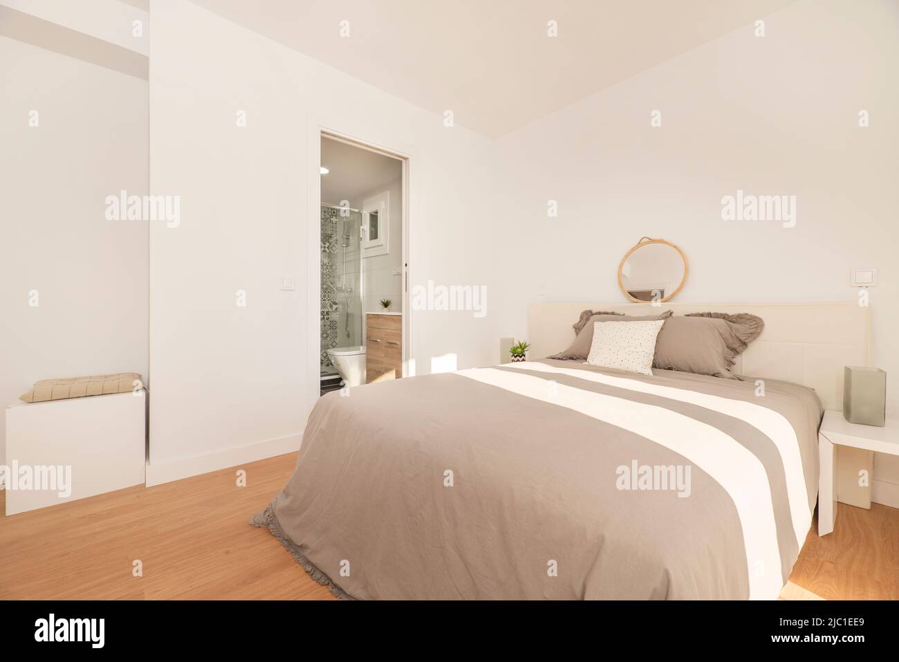 Bedroom with double bed with gray bedspread, white furniture and headboard, access to an en-suite toilet and light wooden floor Stock Photo