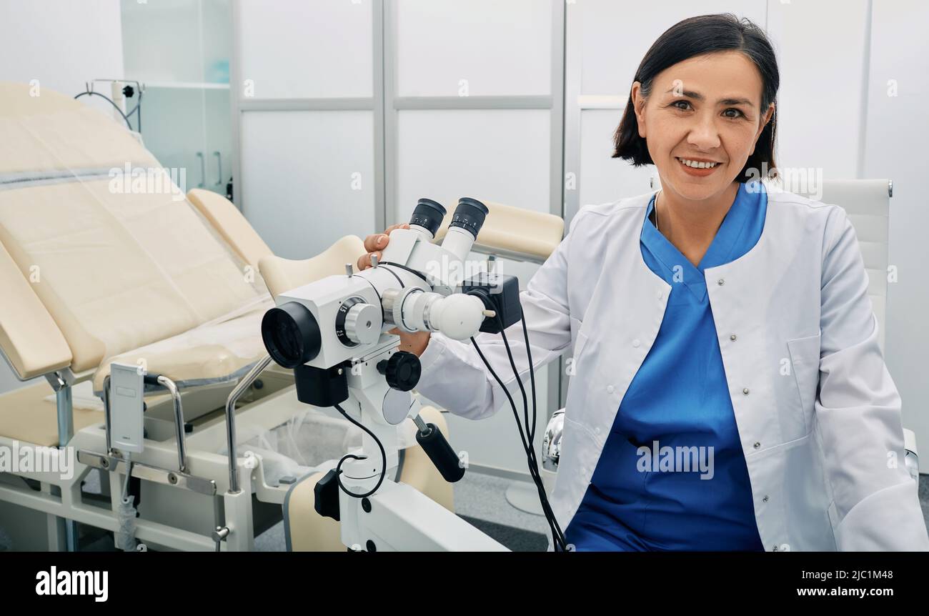Portrait of gynecologist woman sitting near colposcope and gynecological chair in her doctor's office. Gynecologist profession Stock Photo