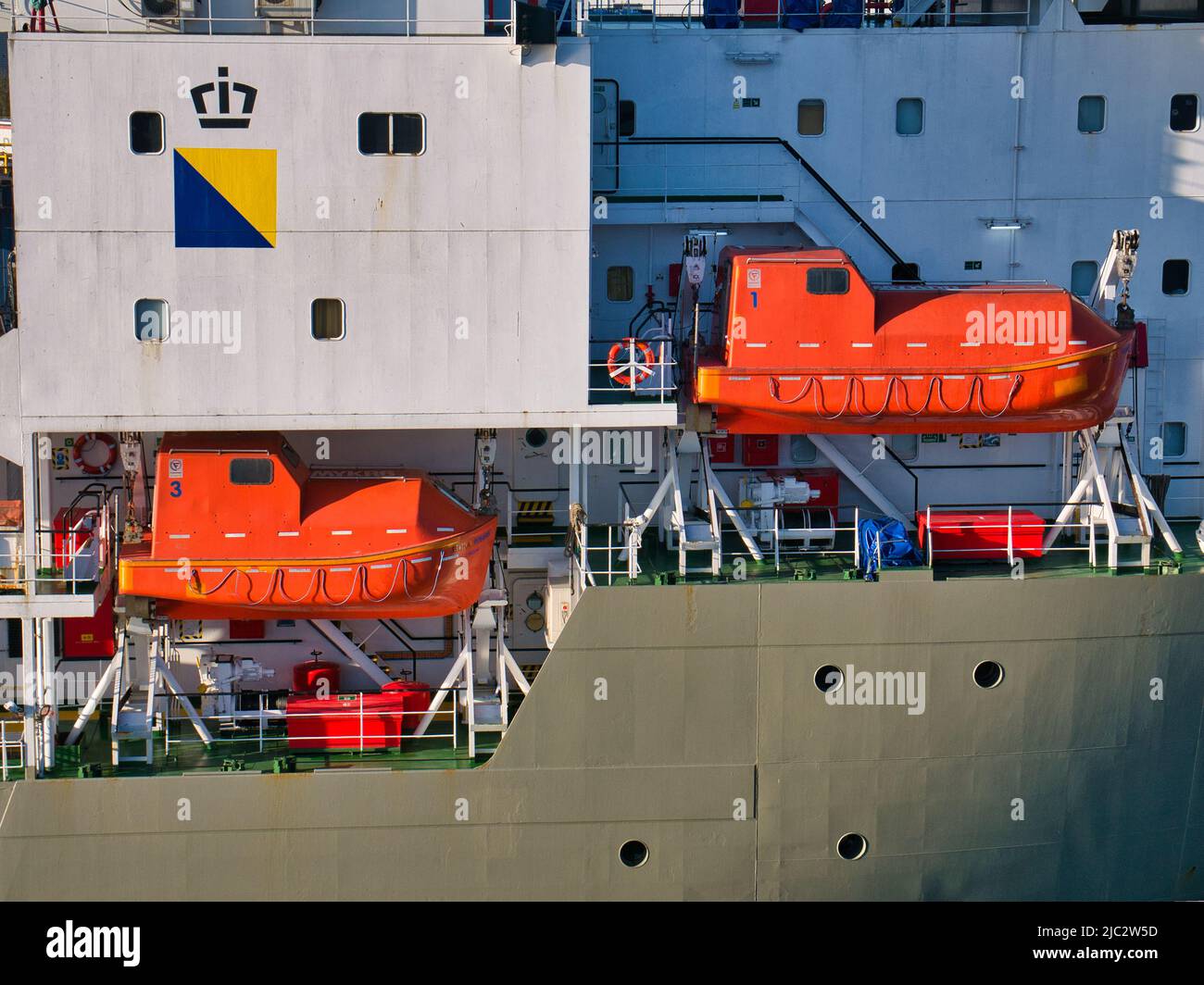 Two orange, high visibility, launch ready marine lifeboats, suspended between white launch davits on a North Sea support vessel in Aberdeen Harbour. Stock Photo