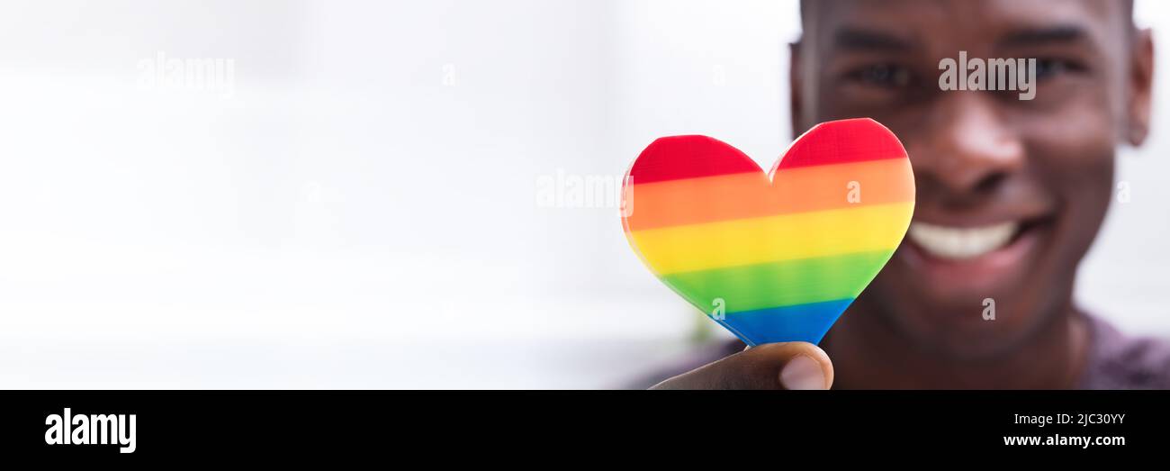 Smiling Man Holding Rainbow Heart In His Hand Against White Background Stock Photo