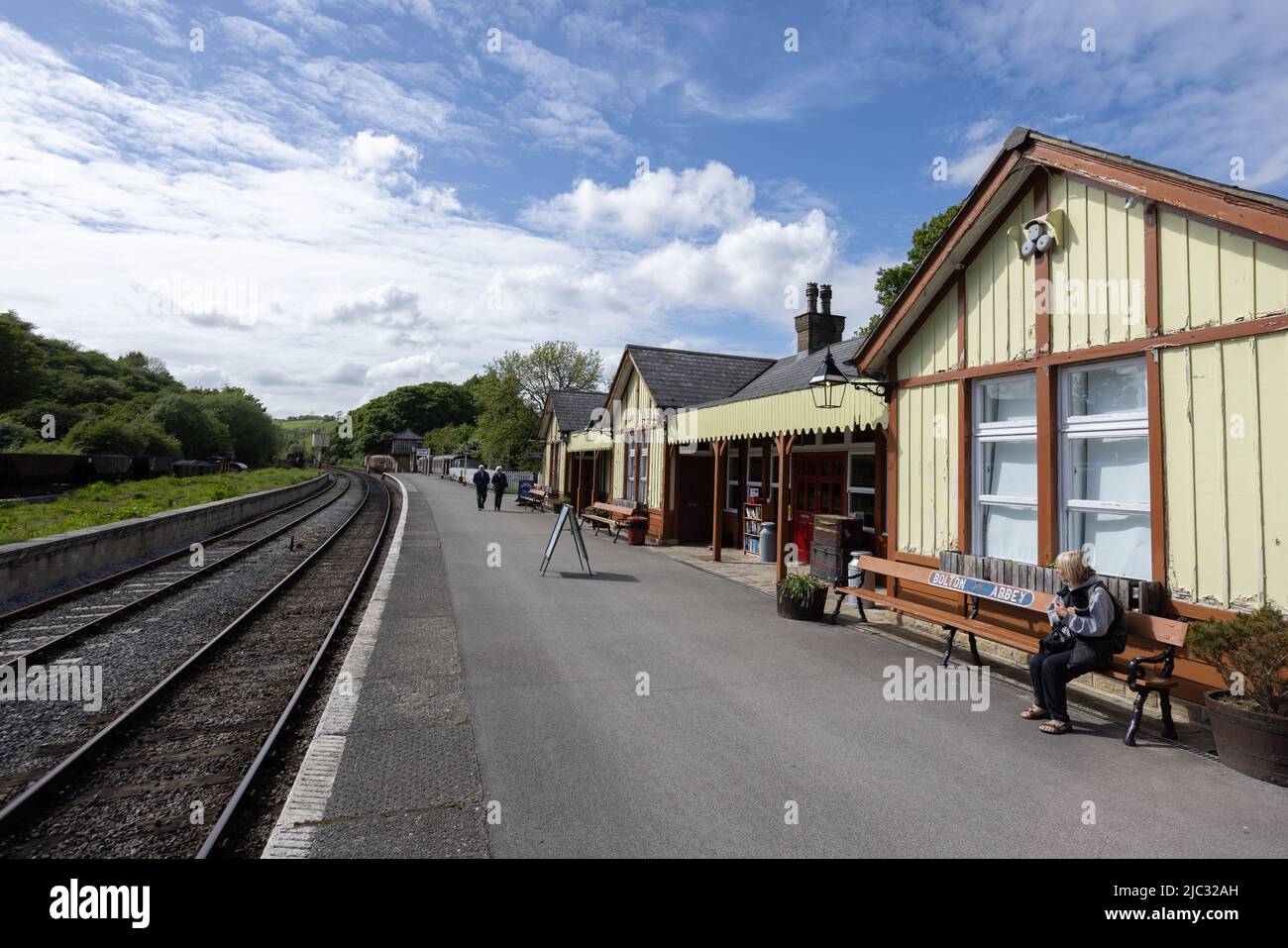 Woman sitting on bench and people walking on platform between colourful yellow wooden train station building and rails at Bolton Abbey railway station Stock Photo