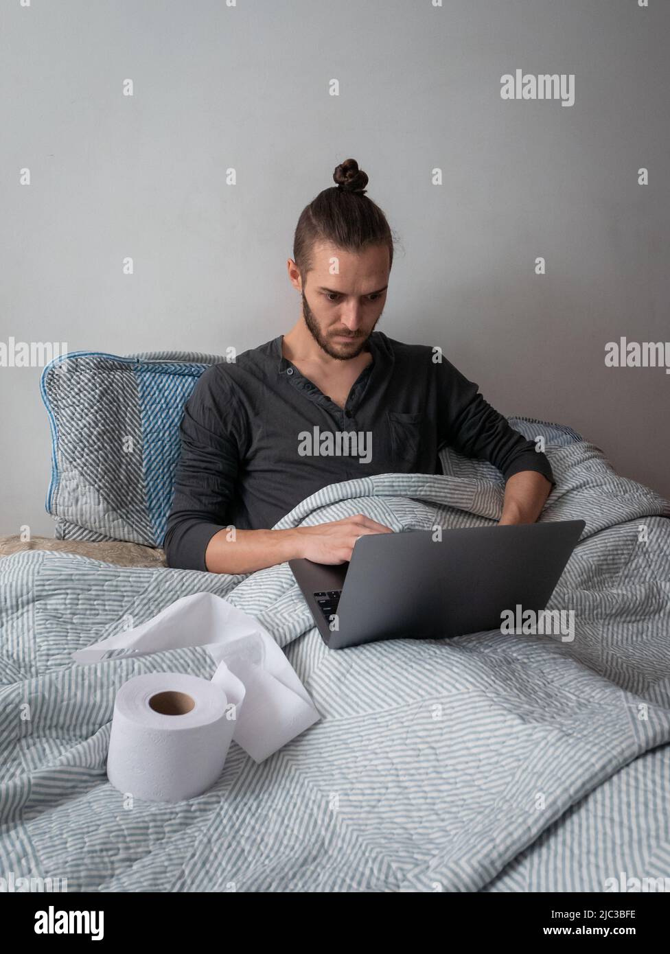 White Man in Bed with a Cold is Working on his Laptop Computer next to a Toilet Paper for his Nose Stock Photo