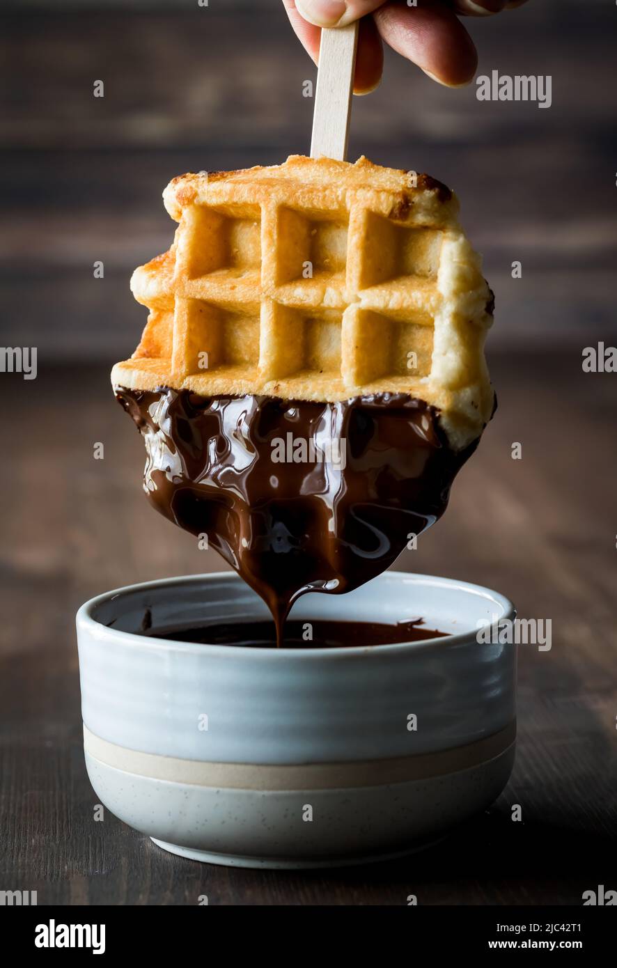 A waffle being dipped into melted chocolate and dripping into a bowl. Stock Photo
