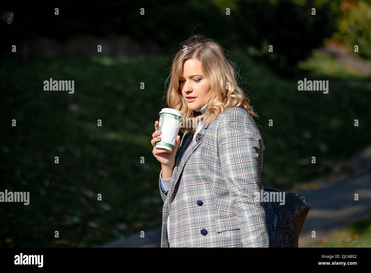 Elegant blonde with long curly hair in light tweed jacket drinking hot coffee from recyclable cup while walking on sunny autumn day in park Stock Photo