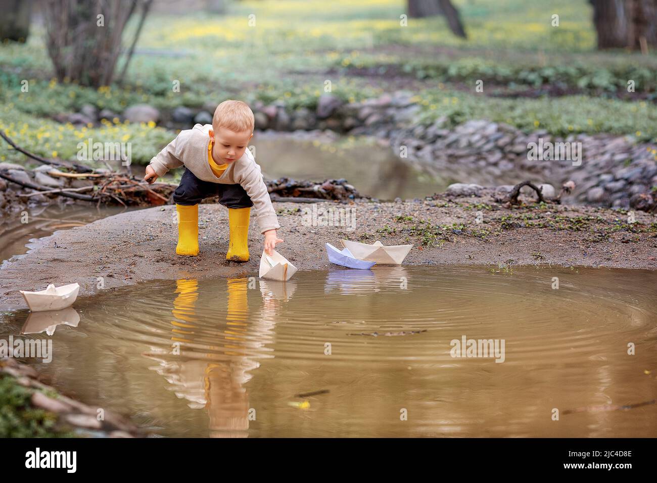 Portrait of adorable little kid boy in rubber boots launch paper boats ...
