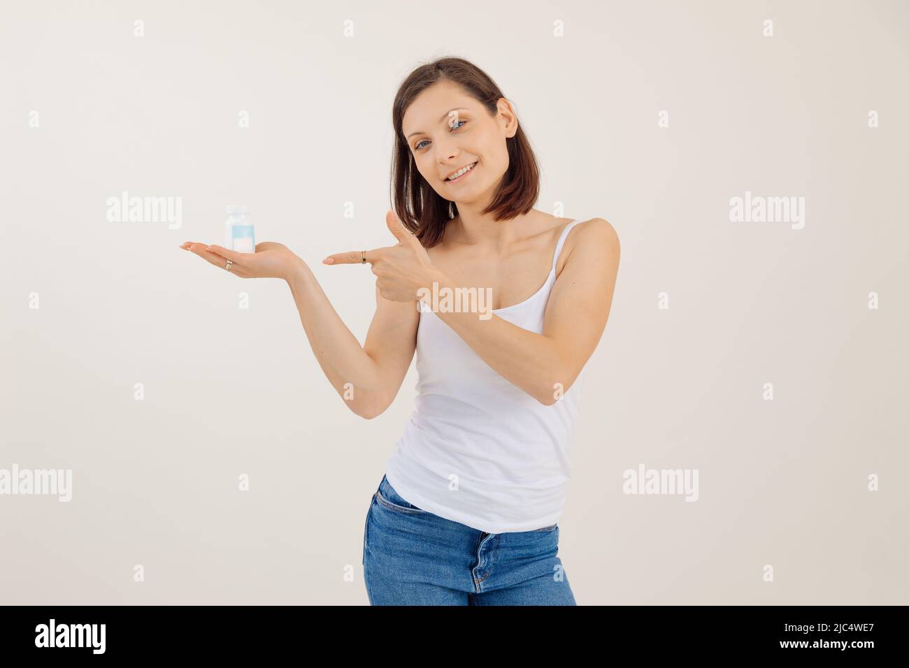 Portrait of young happy woman advertising useful vitamins for women, white background. Holding in hand and pointing at jar of pills, disease treatment Stock Photo