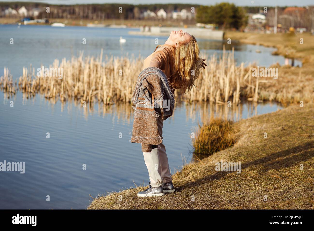Happy beautiful blonde woman with open arms standing near river bank alone on cool sunny day, relaxing, rejoicing and enjoying freedom. Outdoors Stock Photo