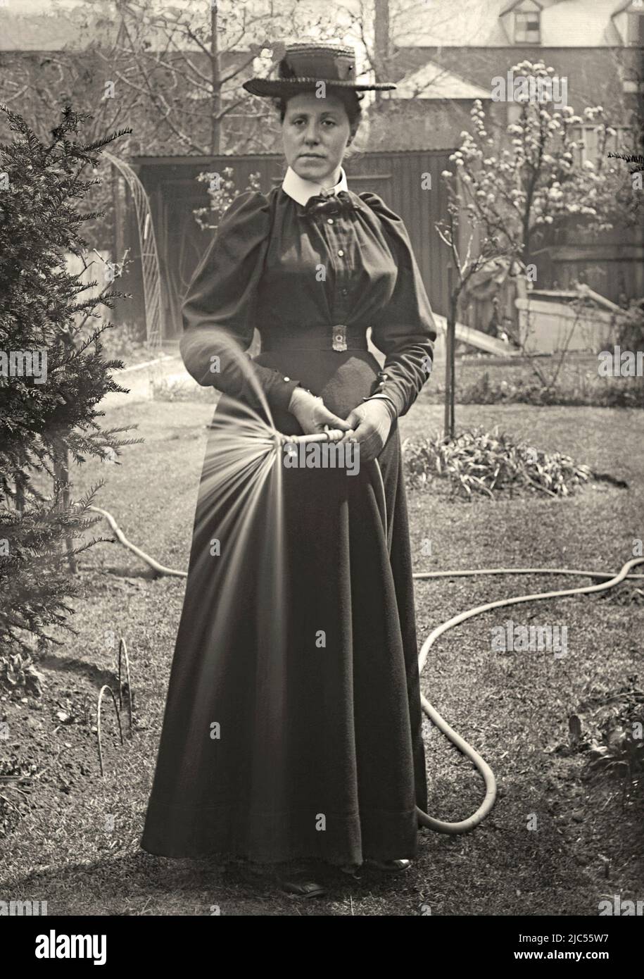 A woman using a hose to water her back garden, UK c. 1900. She wears smart clothing, including a hat and gloves, for the task. This is from an old Victorian glass negative – a vintage 1800s/1900s photograph. Stock Photo