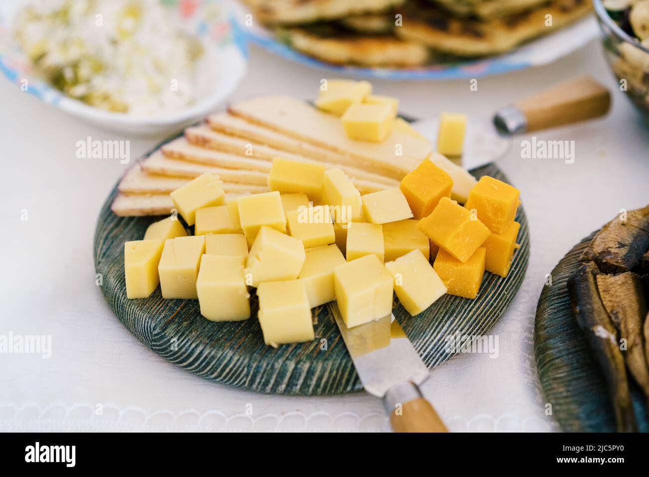 High angle view of cheese, knife, and salad dishes on table. Cheese platter with different sorts of cheese. Stock Photo