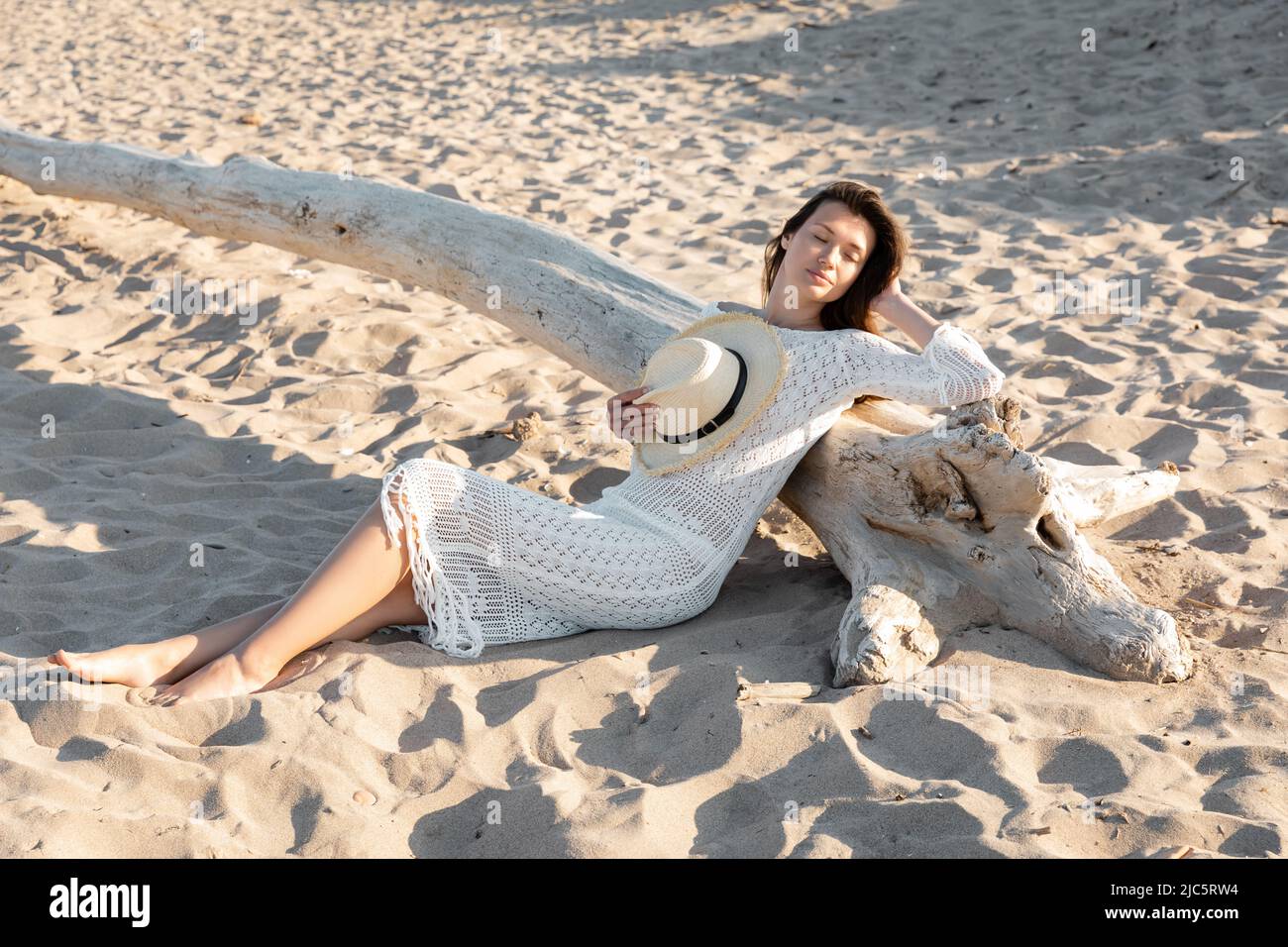 Young woman in dress holding sun hat while resting near wooden log on ...