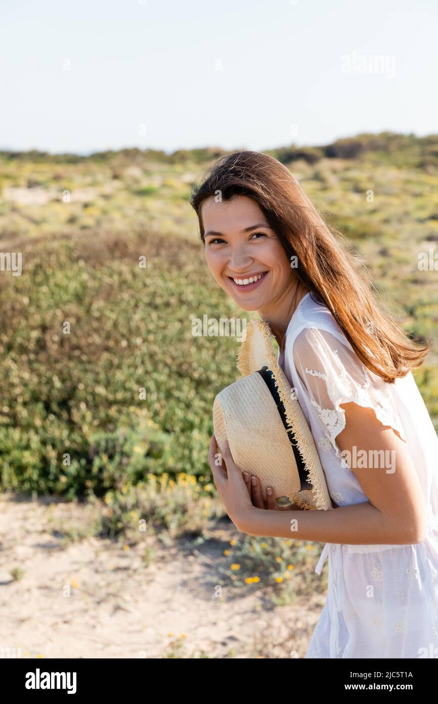 Positive woman in summer dress holding straw hat on blurred beach Stock ...