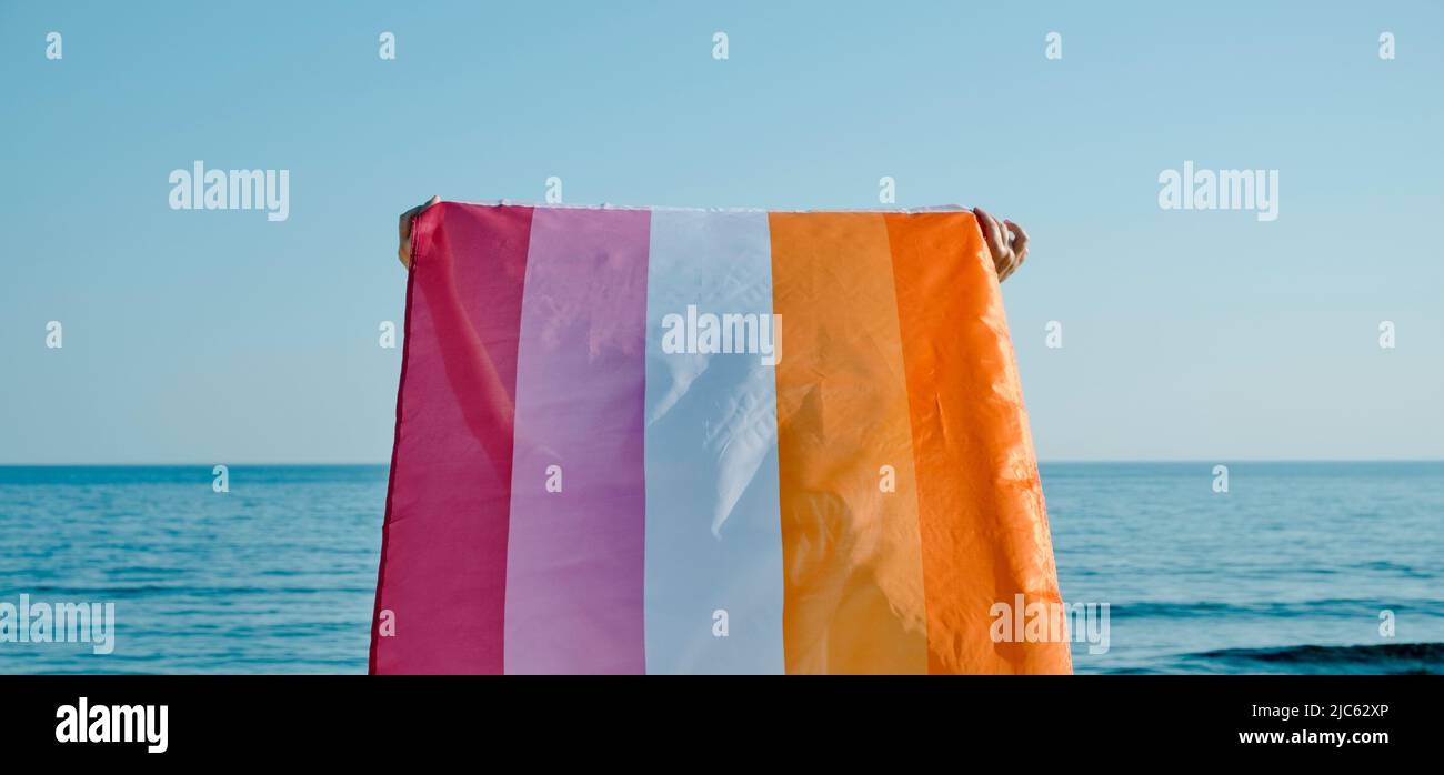 closeup of a woman, seen from behind, holding a lesbian pride flag on the air above her head facing the sea in a sunny day Stock Photo