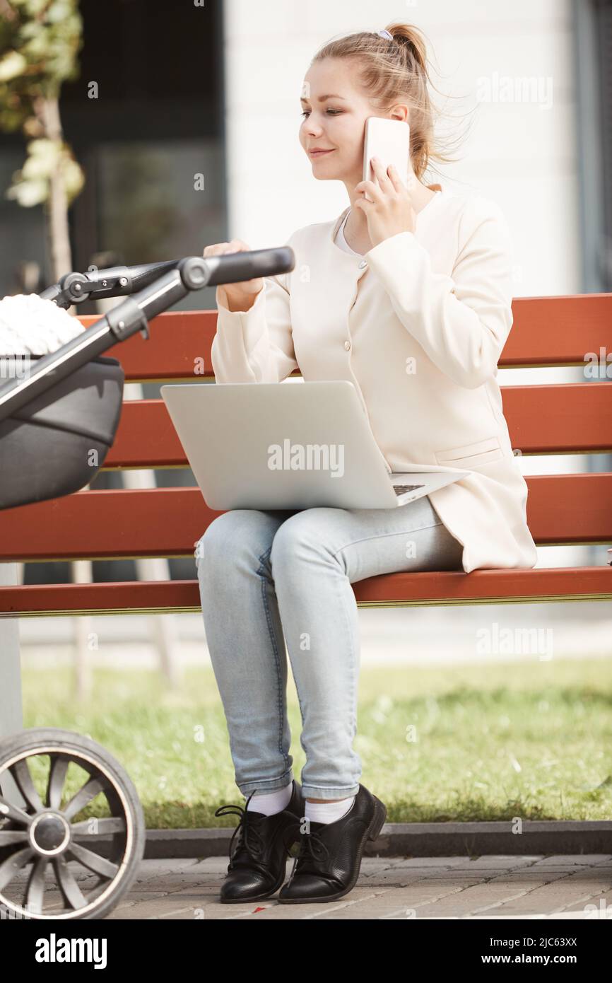 Young mother in white coat working outside on her smartphone and laptop while baby sleeping in stroller Stock Photo