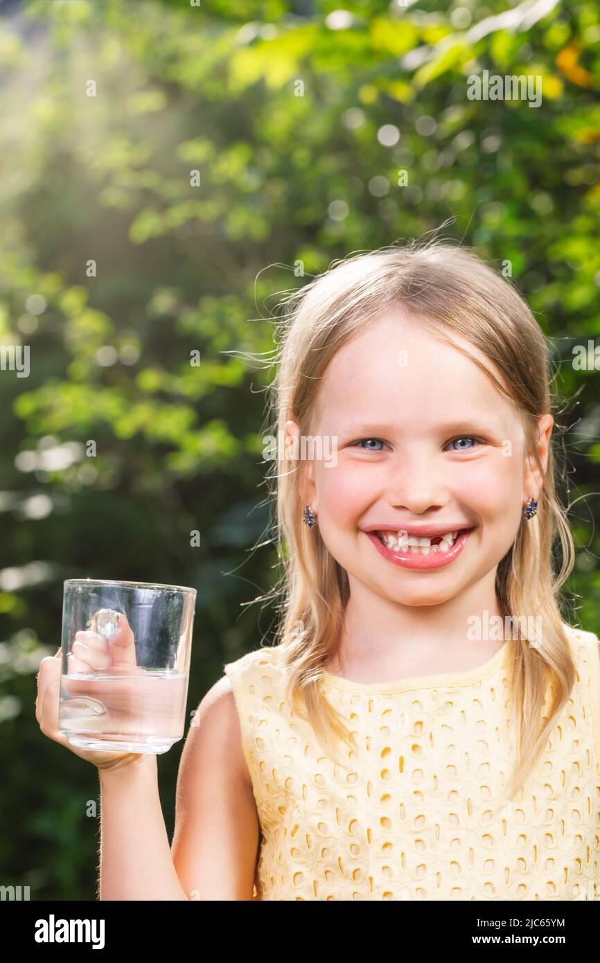 Happy little girl wearing yellow dress holding glass cup of water in a summer garden - healthy lifestyle concept Stock Photo