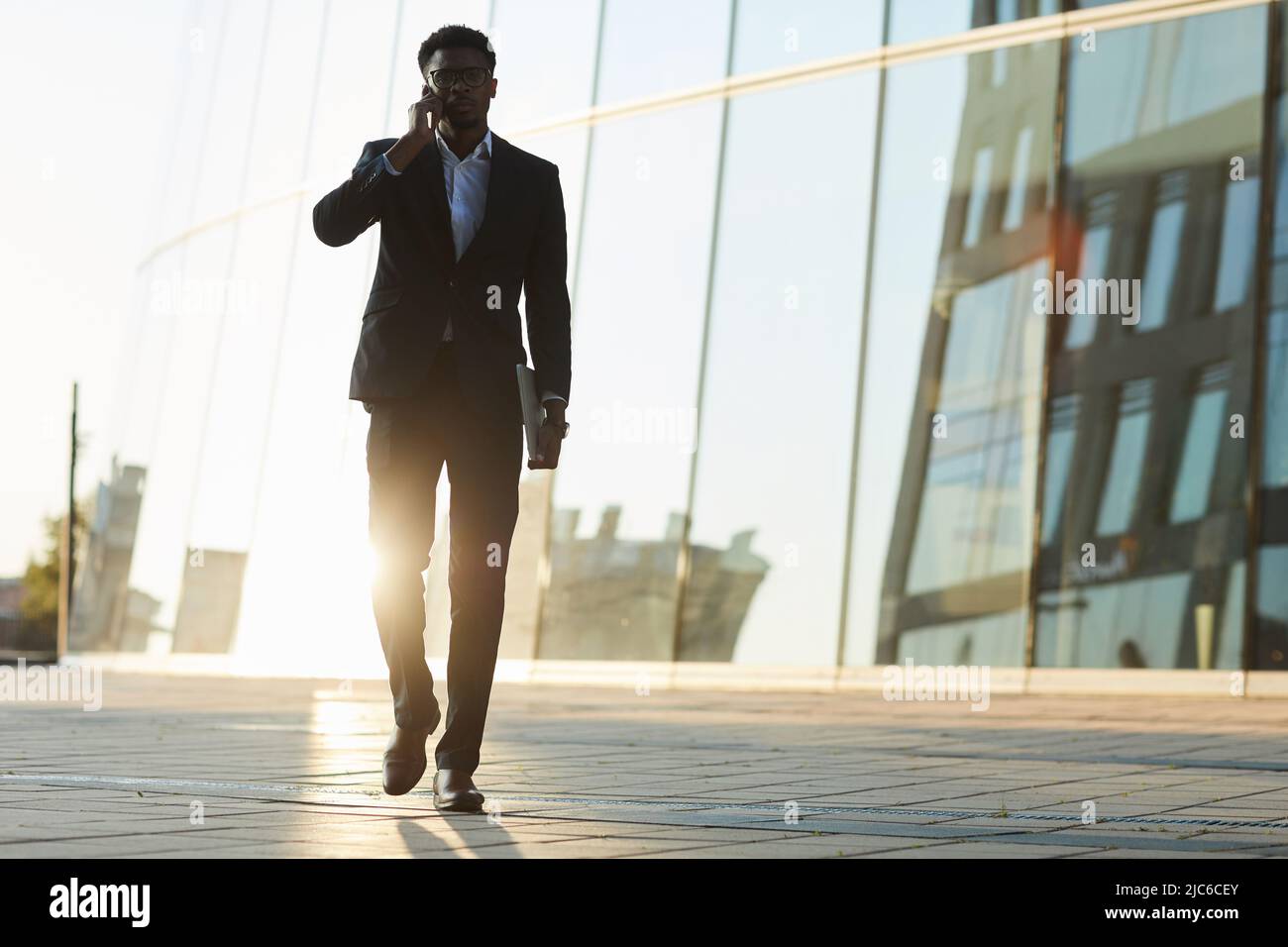 Confident young African-American businessman in formal suit walking over business district and talking by cellphone, he working on move Stock Photo