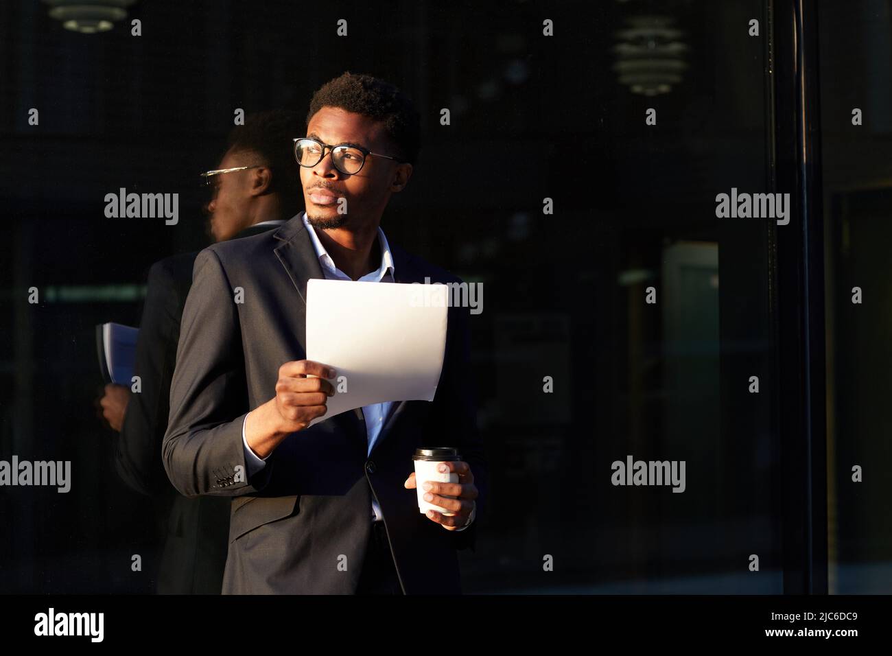 Serious pensive young Afro-American man with beard standing by office building and drinking coffee while reading papers Stock Photo