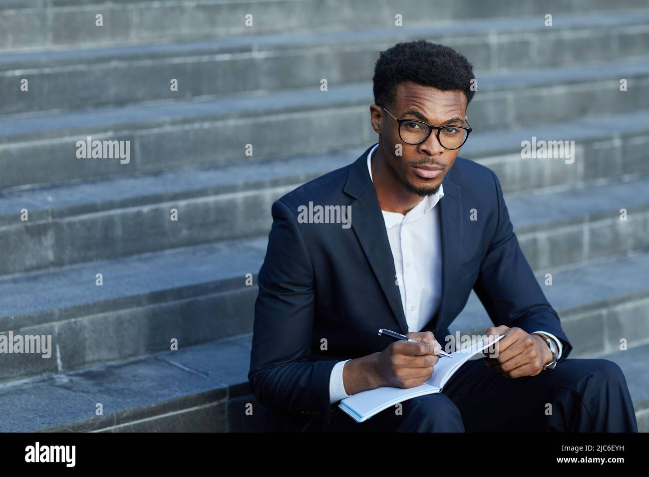 Serious thoughtful young Afro-American business school student with furrowing forehead sitting on staircase and making note in notepad Stock Photo