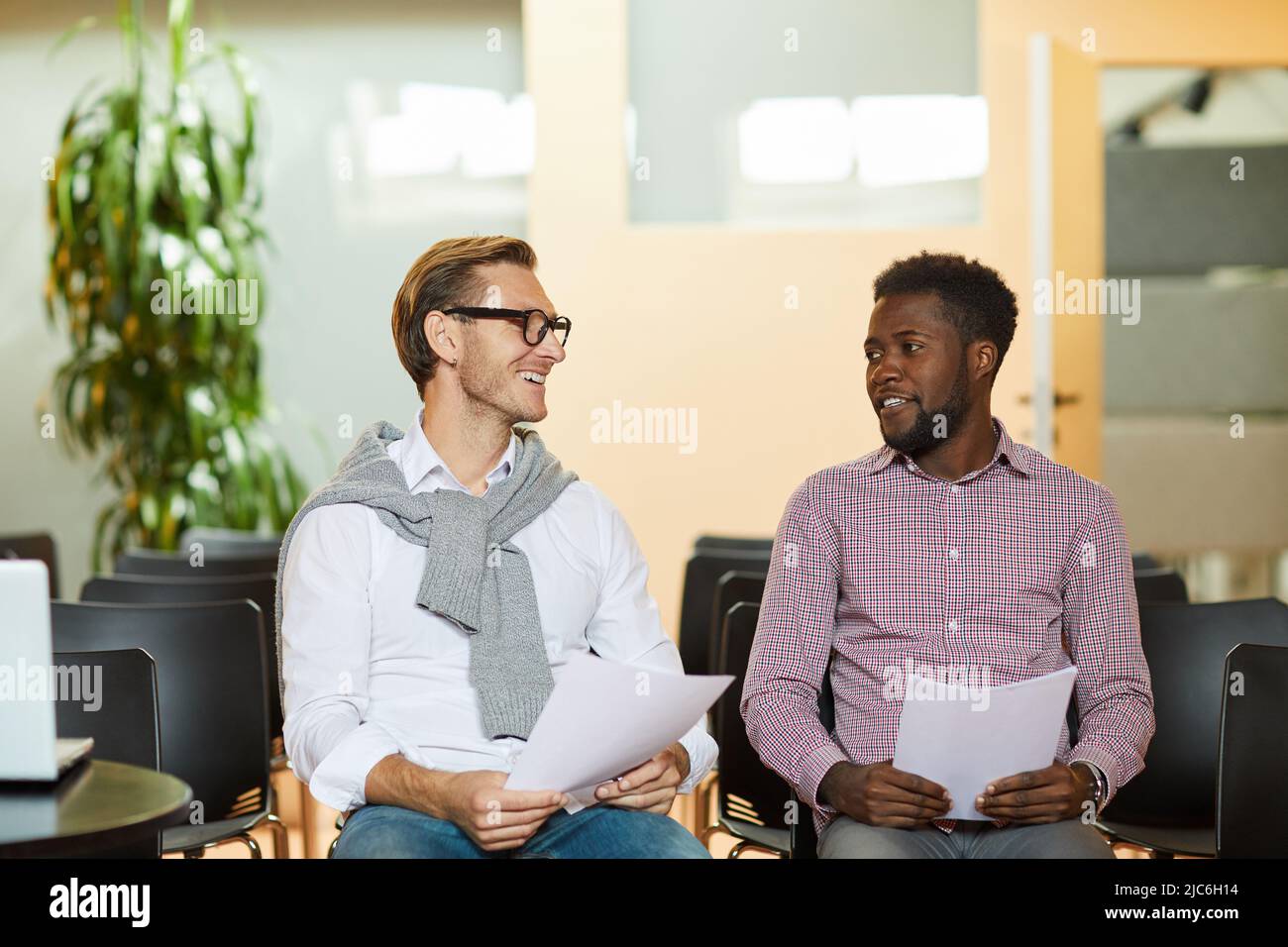 Smiling young multi-ethnic specialists sitting on chairs in auditorium and sharing ideas of marketing strategy Stock Photo