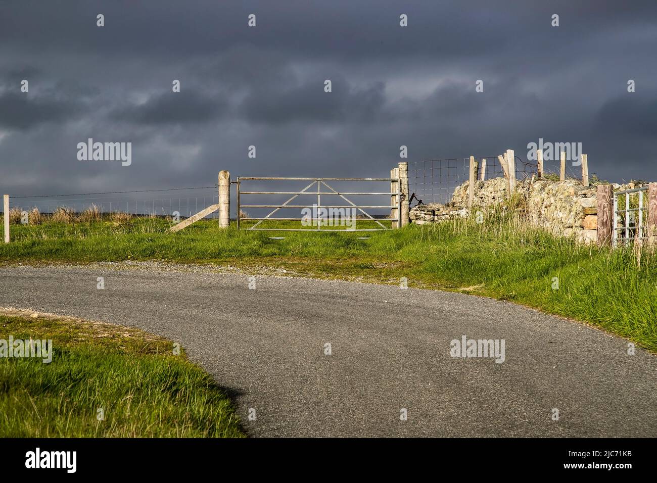 Five bar steel gate on the corner of a country road and entrance to farmland with dark gathering clouds beyond heralding an approaching storm Stock Photo