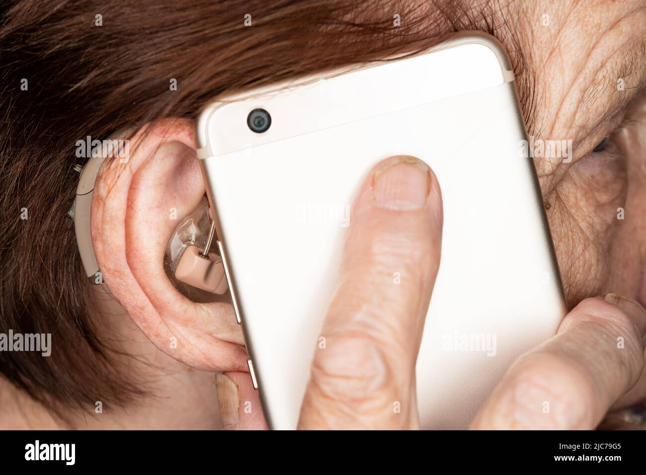 Elderly senior woman holding mobile phone next to her ear with hearing aid device, closeup detail Stock Photo