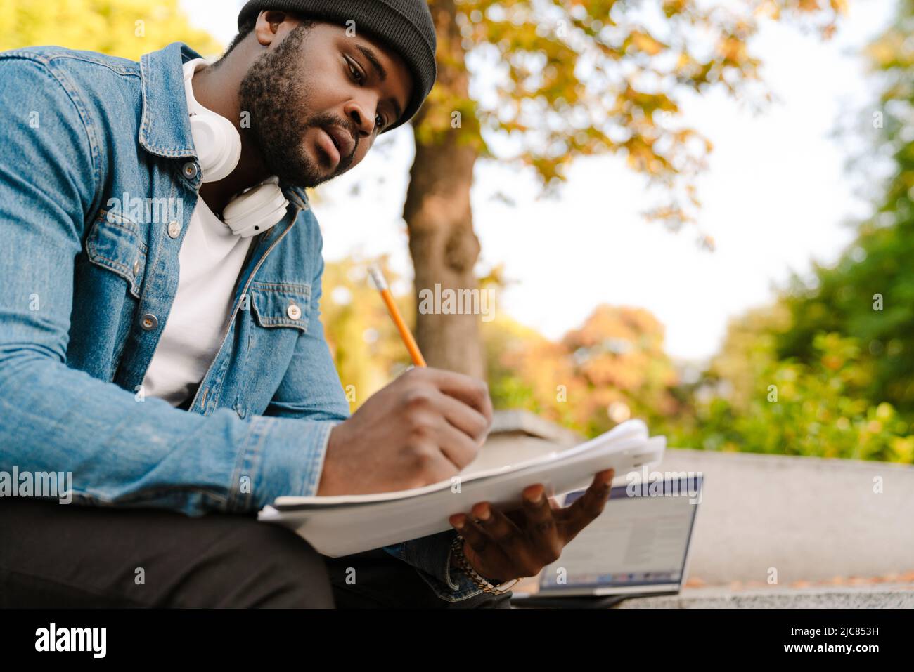 Black man writing down notes while working with laptop on bench ...