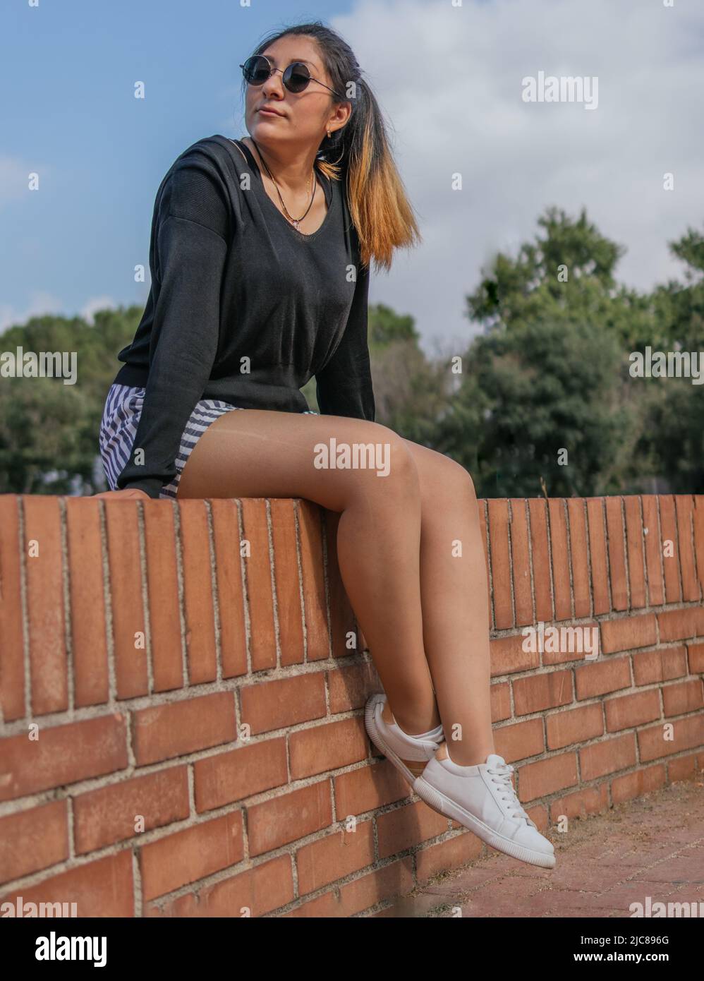 A beautiful lady sitting on a brick railing is swinging her legs and looking aside in a town square Stock Photo