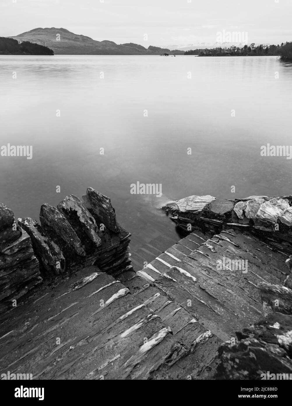 Black and white Stunning landscape image of Loch Lomond and snowcapped mountain range in distance viewed from small village of Luss Stock Photo