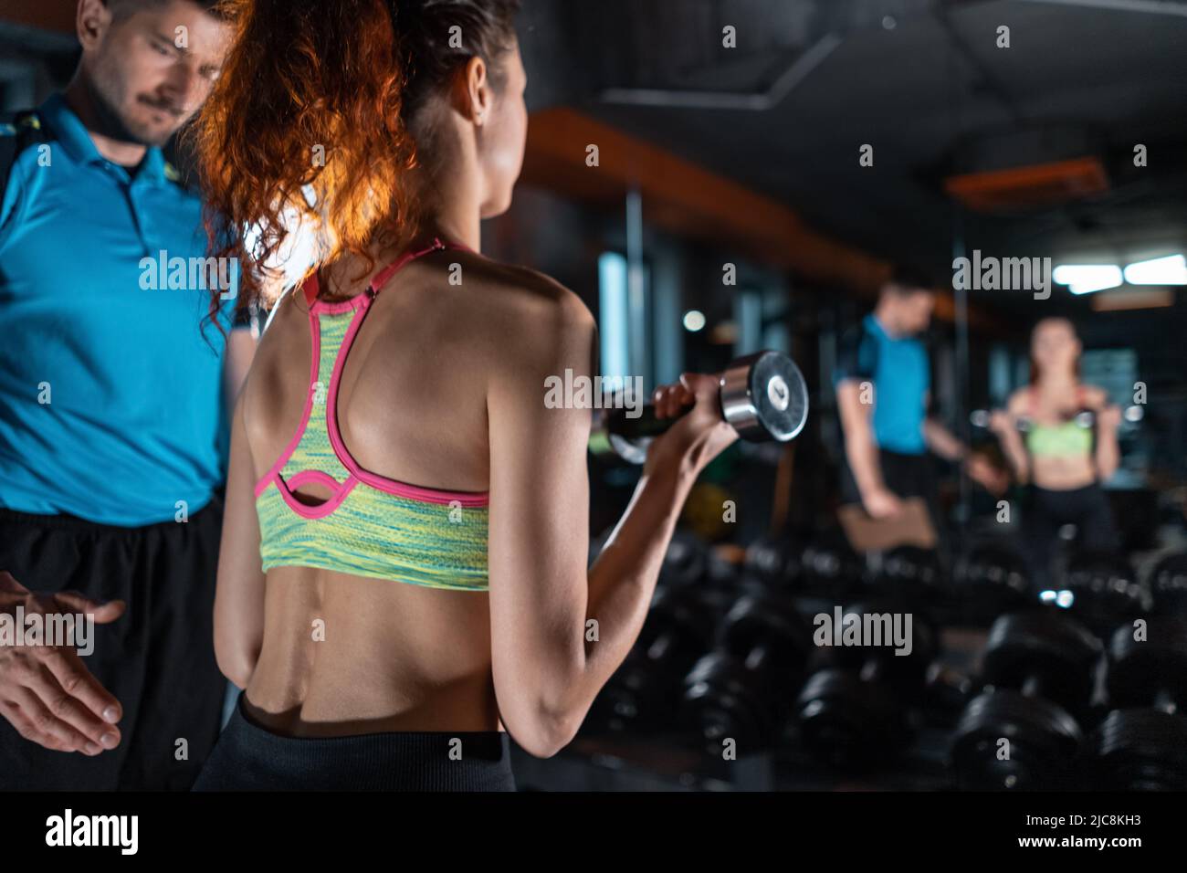 back view of female exercising in gym with dumbbell with personal trainer Stock Photo