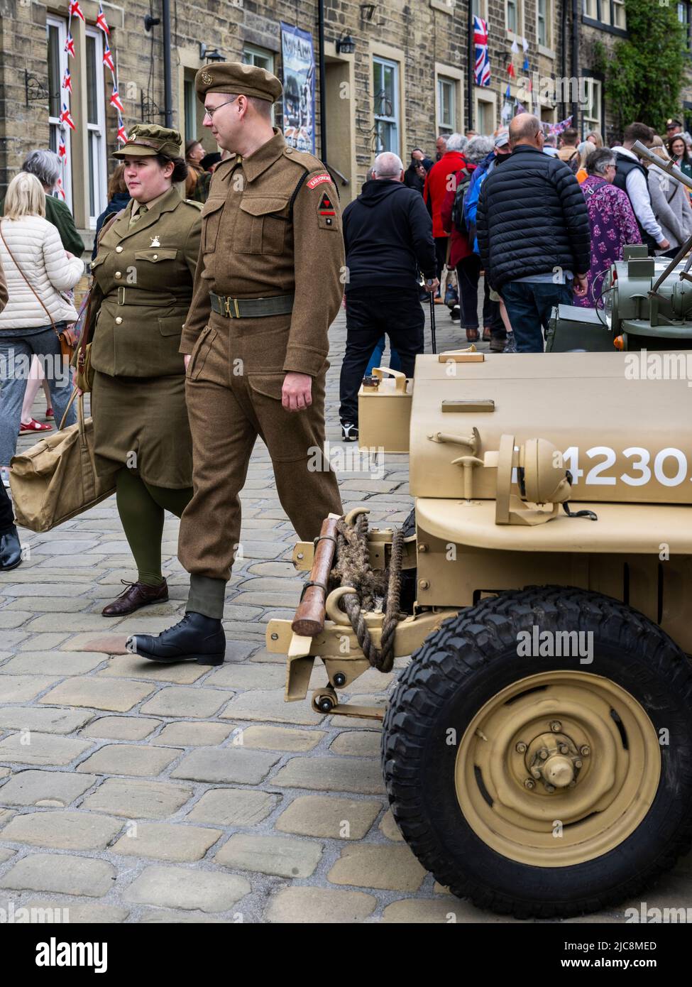 Haworth 1940's weekend (male dressed as soldier passing old Jeep, female in khaki, crowds on busy scenic Main Street) - West Yorkshire, England, UK. Stock Photo