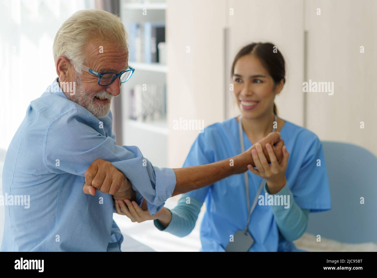 Senior man physiotherapy exercises with Asian woman caregiver or nurse physiotherapist in uniform helping aged male patient at home. Stock Photo