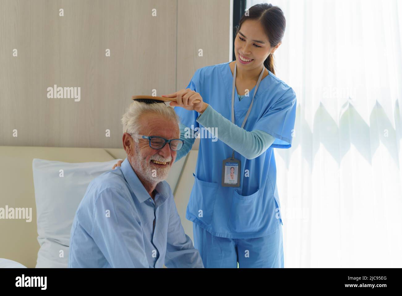 Young Asian woman nurse helping brushing hair to disabled elderly man in bed at retirement home. Millennial caregiver assisting handicapped senior pat Stock Photo