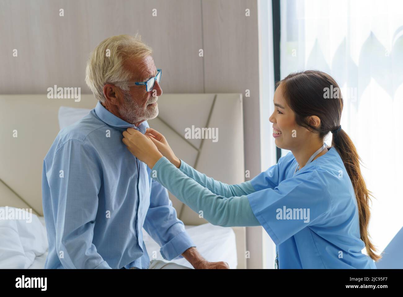 Young Asian woman nurse helping get dress to disabled elderly man in bed at retirement home. Millennial caregiver assisting handicapped senior patient Stock Photo