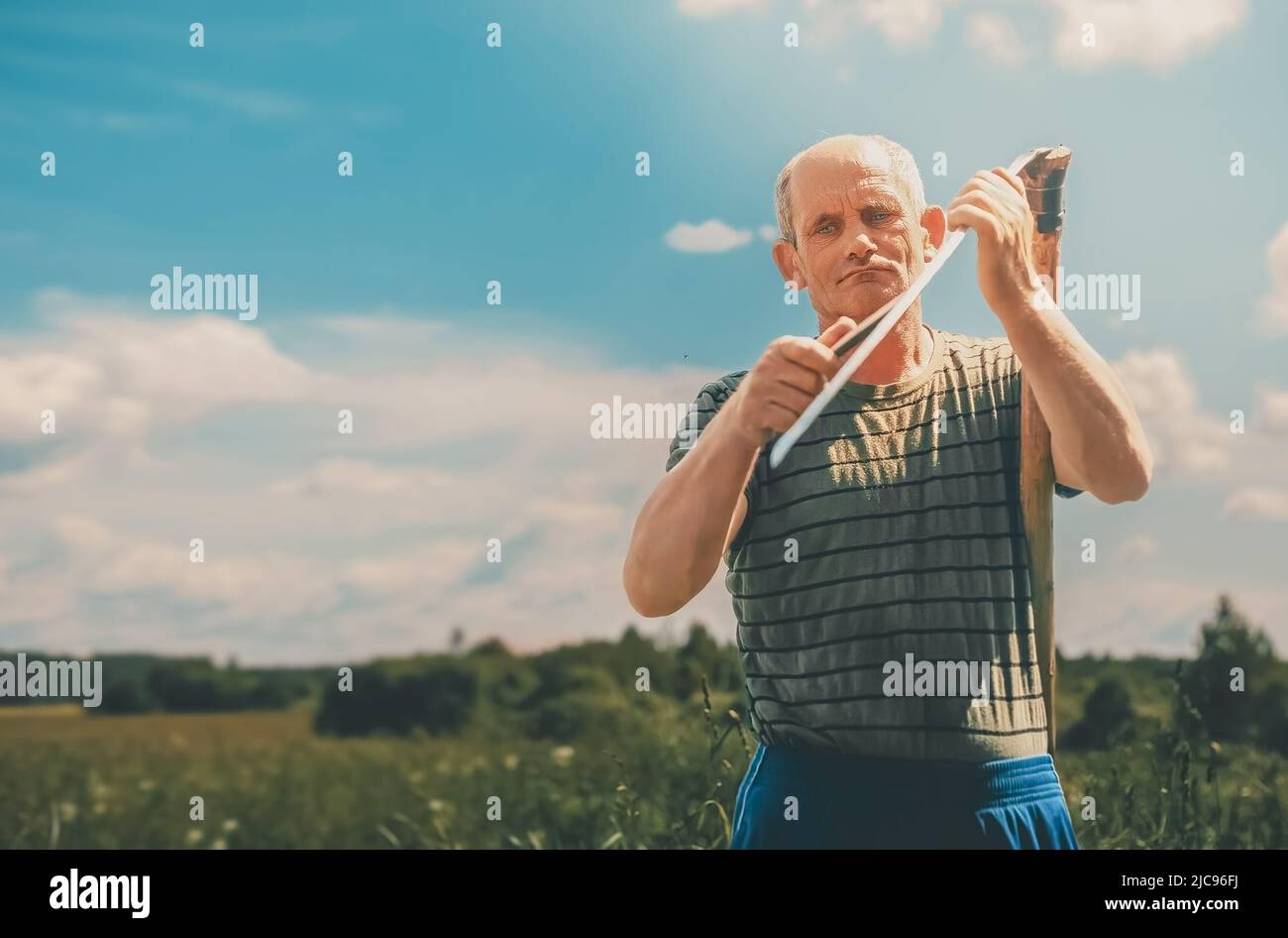 A man grinds an old scythe to mow grass in a meadow in summer Stock Photo