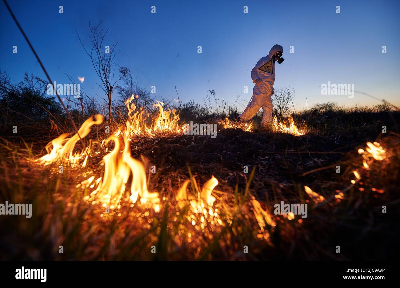 Firefighter ecologist fighting fire in field at night. Man in protective suit and gas mask near burning grass with smoke, holding warning sign with skull and crossbones. Natural disaster concept. Stock Photo