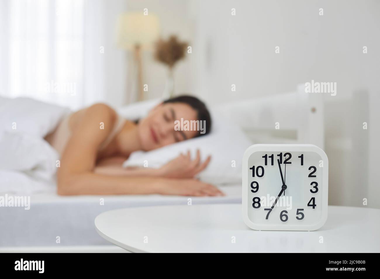 Alarm clock set for 7 AM on bedside table, with happy woman sleeping on bed in background Stock Photo