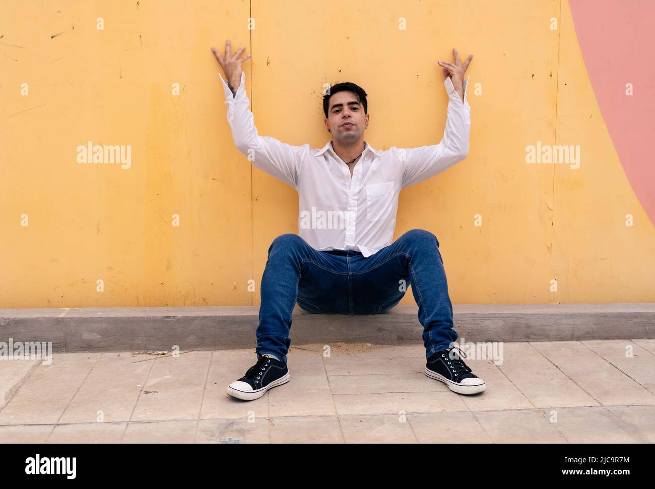 Young latino guy sitting and leaning against a yellow wall with his arms open up. Modern clothing. Stock Photo