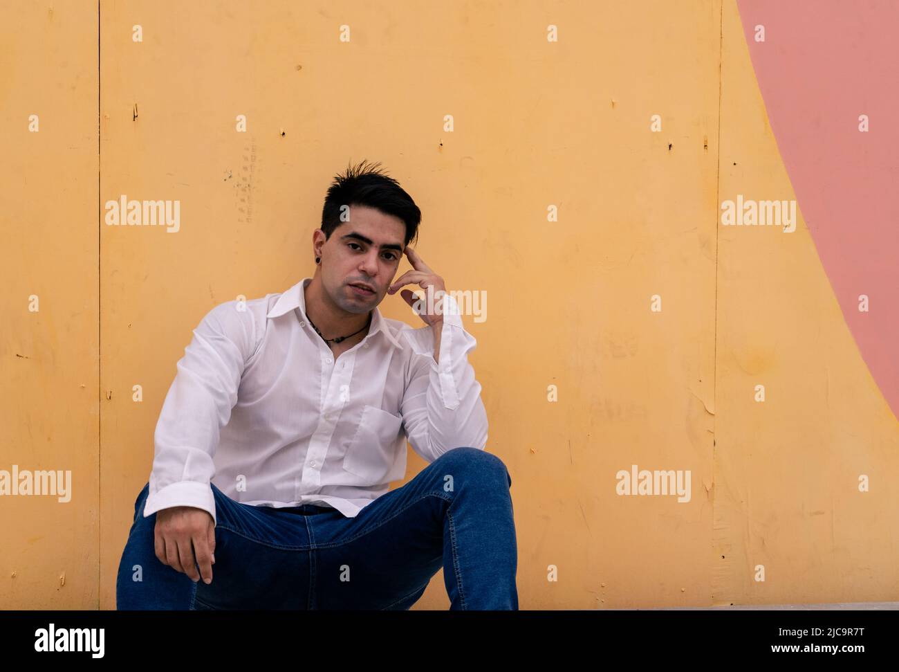 Young latino guy sitting and leaning against a yellow wall in a pensive attitude. Modern clothing. Stock Photo