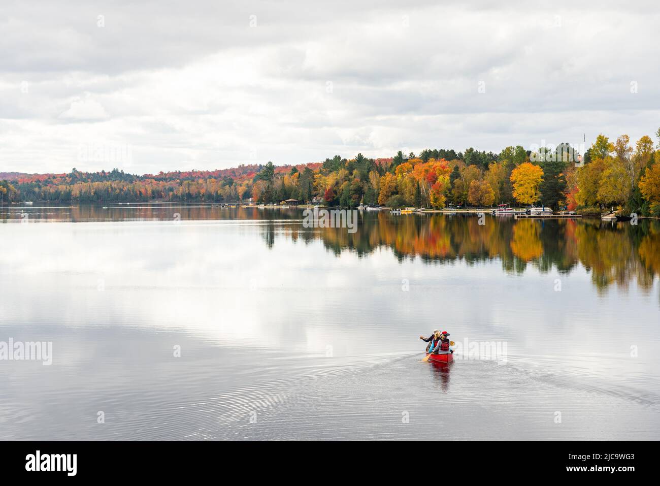 People canoeing on a beautiful lake surrounded by forest at the peak of fall foliage on a cloudy autumn day Stock Photo