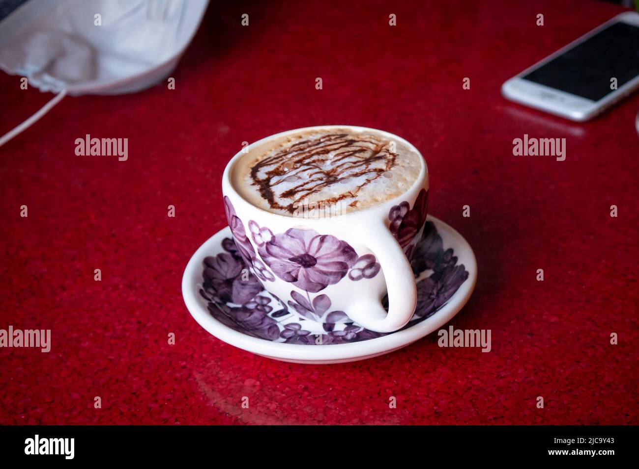 Capuccino Coffee with Chocolate Drizzles in a Purple Flowered Cup by a Smartphone and a Face Mask Against a Red Table Background Stock Photo