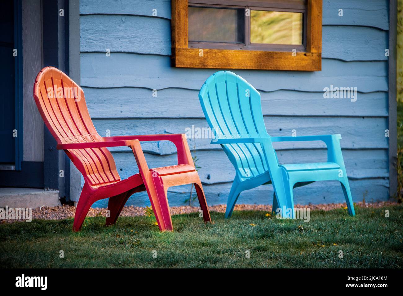 Close-up of red and turquoise arondiack chairs sitting outside blue wooden cabin door on grass. Stock Photo