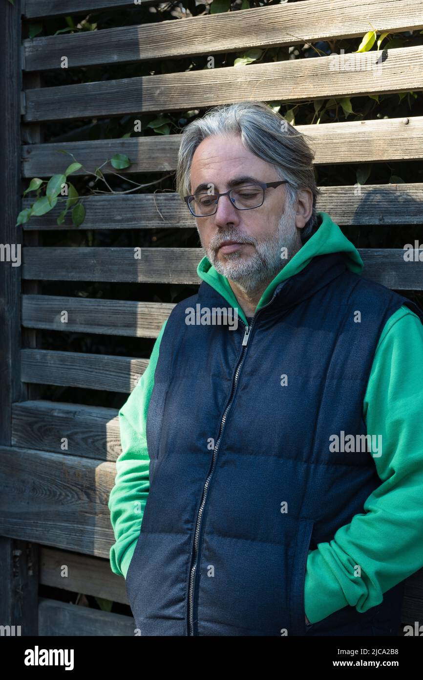 Mature man with grey hair and glasses leaning on the wooden fence, thoughtfully looking down with his hands in his pockets. Stock Photo