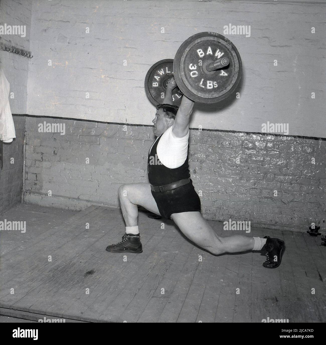1957, historical, weight-lifting, inside a gym on a wooden board, a male weightlifter training, lifting the barbell doing the clean-and-jerk, Stockport, Manchester, England, UK. On the barbell is 60lbs. The clean-and-jerk is one of the two lifts in competitive or Olympic-style weightlifting. He is wearing weightlifting shoes and the traditional leather weightlifting belt. On the barbell, a logo with the initials BAWLA, the British Amateur Weightlifting Association, named as such in 1911 after being founded in 1910 as the BWLA. Stock Photo