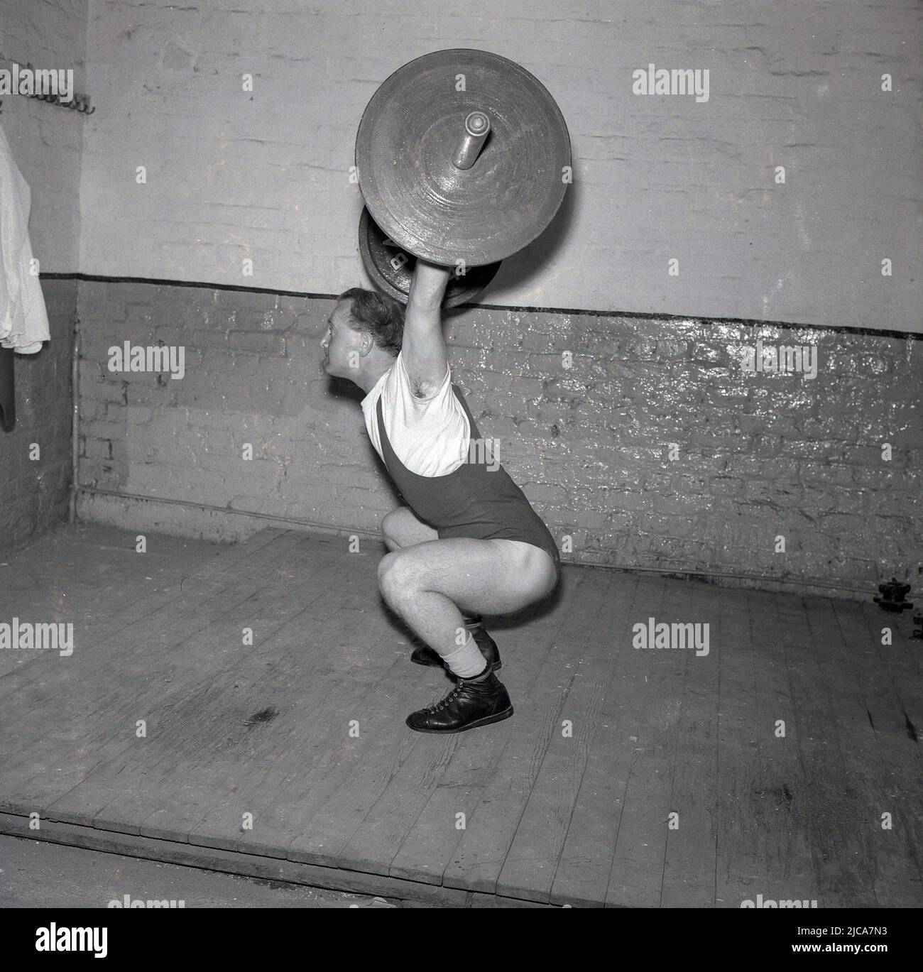 1957, historical, weight-lifting, inside a gym on a wooden board, a male weightlifter training, doing the snatch, Stockport, England, UK. The snatch is one of the two lifts in competitive or Olympic-style weightlifting. He is wearing weightlifting shoes, but is not wearing the traditional wide leather weightlifting belt. Stock Photo