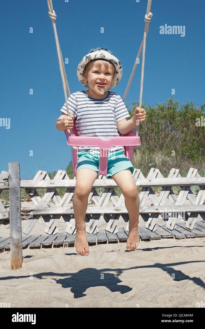 The little boy sways in the beach swing. The child is very cheerful, and naughty he shows his tongue Stock Photo